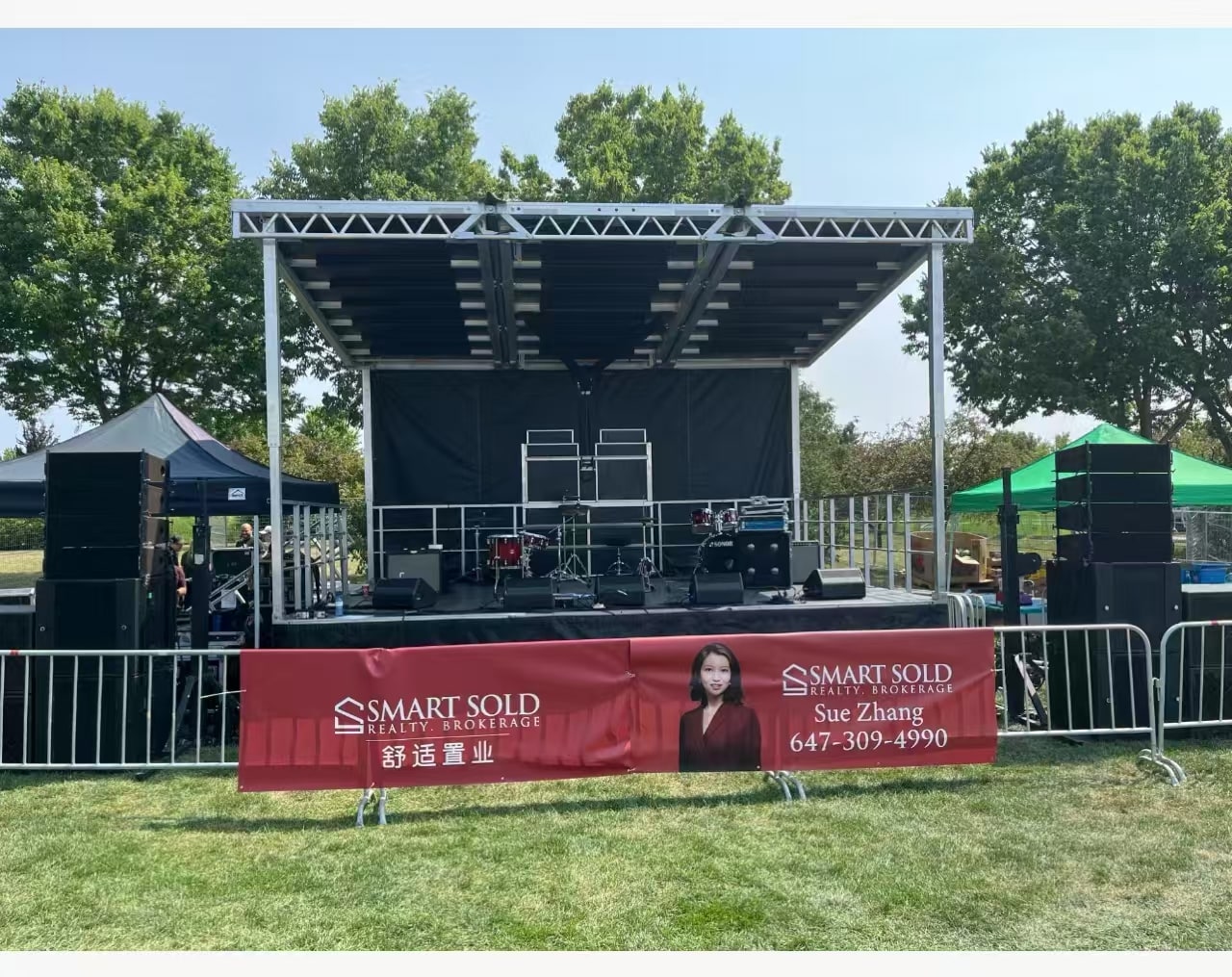 An outdoor stage with a Smart Sold Realty banner featuring Sue Zhang and her business phone number.